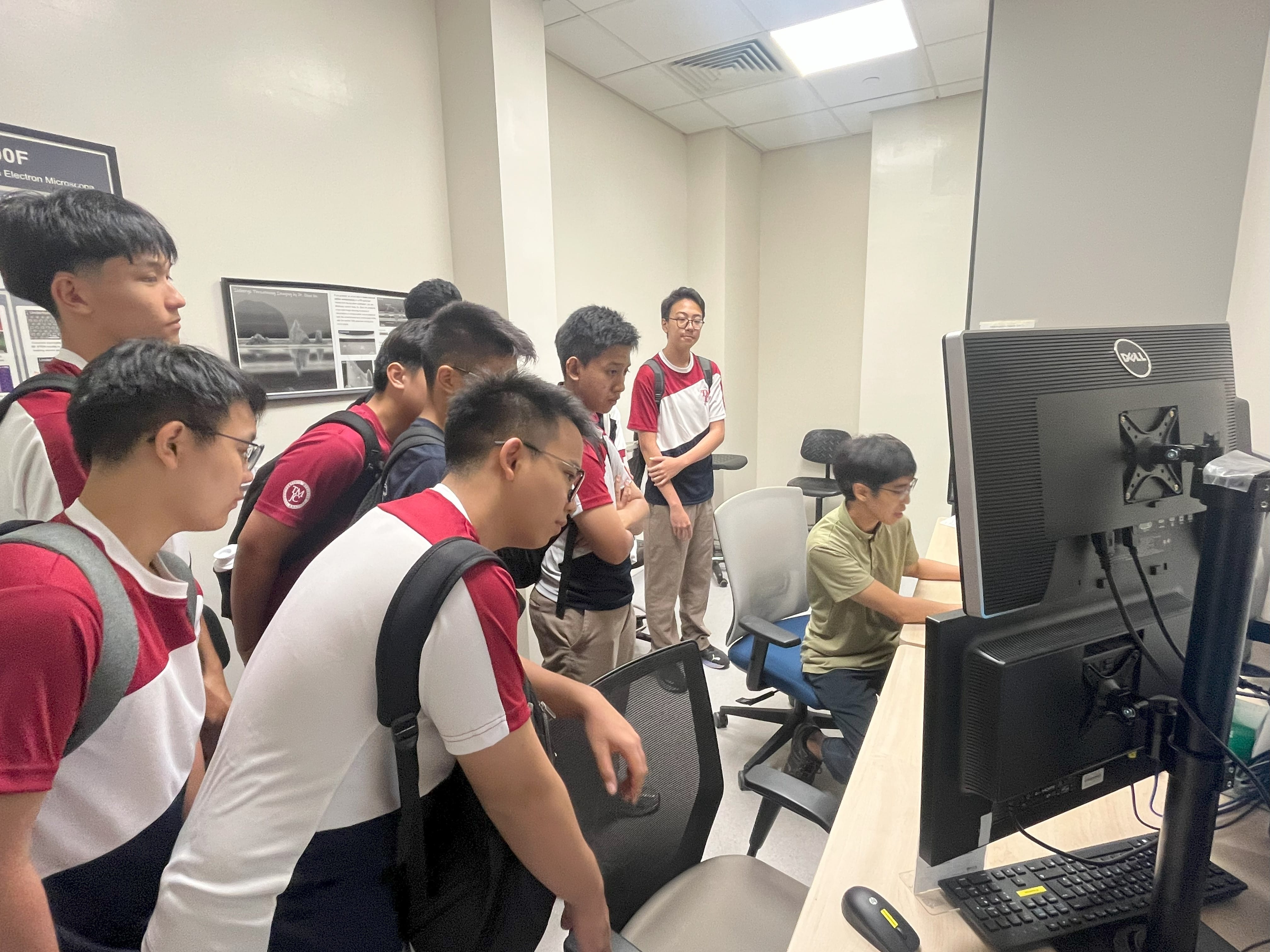 A group of male students in school uniforms crowd around a person seated at a computer workstation, watching a demonstration. A research poster is visible on the wall behind them.